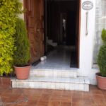 Fotografía Entrada Sacramento Domicilio Particular donde se puede apreciar dos escalones de azulejo blanco que dirigen a la puerta de entrada que es de madera la cual está abierta y deja ver un poco el pasillo y donde también se encuentran a la entrada dos plantas en una maceta grande de barro cada una y un arbusto recargado en la pared del lado izquierdo, en el marco derecho de la puerta se encuentra un letrero de cerámica con la leyenda "La casa de los abuelos" y debajo de este un barandal de apoyo para personas con discapacidad