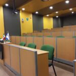 Se aprecian 6 filas de mesas de trabajo tipo auditorio color café con sillas verdes y un proyector en la parte de arriba en una sala con paredes pintadas con franjas amarillas y grises.
