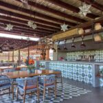 Interior de un restaurante al aire libre. Techo de palapa con luces en forma de estrella colgando. Mesas de madera con sillas a juego, con cojines azules y blancos, sobre un piso de baldosas geométricas. Una barra de servicio con azulejos verdes y blancos en patrón de diamantes cuenta con lámparas industriales negras colgando sobre ella.