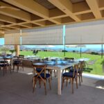 Terraza de campo de golf al aire libre con mesas de madera y sillas con cojines azules, bajo techo beige. La vista da a un césped y el mar, con colinas en la distancia.