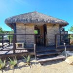 Vista de una cabaña con techo de paja, construido sobre una plataforma de madera con barandilla, rodeado de plantas. La estructura se asienta en un paisaje árido.