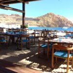 club con mesas de madera y sillas con cojines azules bajo un techo rústico de palapa. Ofrece una vista panorámica del mar con barcos y una colina rocosa al fondo