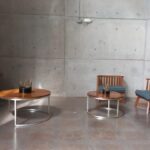 Área de espera con dos mesas redondas de madera de distinto tamaño y 2 sillas de madera con cojín azul y una lámpara de techo en color gris