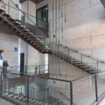 Escalera interior con barandales de cristal, que conecta varios pisos de el edificio. Con un adorno colgante del techo con elementos de metal y las paredes de concreto.