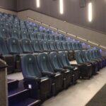 Sala de cine con asientos de color azul oscuro dispuestos en filas escalonadas para una visualización óptima. Las luces de pared proporcionan una iluminación ambiental suave y los pasillos están marcados con luces en los escalones.