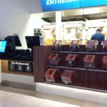 Mostrador en un cine con una fila preferencial para discapacitados, embarazadas y personas mayores. La pantalla muestra un menú digital y hay cajas de palomitas de maíz al fondo y otras golosinas disponibles para la compra al frente