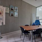 Sala de juntas, con una mesa rectangular y 7 sillas azules, en la pared de concreto colgando a modo de decoración 4 cuadros y a su izquierda una pancarta decorativa.