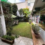 Jardín con césped verde y una pequeña mesa con sillas. Hay varias plantas en macetas junto a la casa a la derecha. El jardín está rodeado por una cerca blanca cubierta de enredaderas a la izquierda. La casa tiene ventanas grandes y está parcialmente cubierta por las sombras del techo extendido.