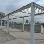 Área exterior con pérgola metálica de forma rectangular con techo blanco en terraza, al fondo un elevador para el acceso