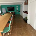 sala de reuniones iluminada con paredes verdes. Hay una larga mesa rectangular de madera en el centro, rodeada por sillas de colores. Un televisor grande está montado en la pared y a su izquierda, hay una pizarra blanca. Hay plantas verdes decorativas colocadas alrededor del espacio.