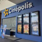 la entrada de un cine Cinépolis. El nombre del cine está en letras grandes en una pared azul oscuro. Debajo de un toldo gris, hay carteles de películas y una puerta cerrada. A la izquierda, hay dos banners promocionales que muestran ofertas especiales o promociones. La estructura tiene acabados modernos con madera y colores oscuros predominantes.