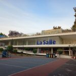 Edificio de dos plantas con rampa al primer piso cuyos muros son de vidrio. Cancha de voleibol y de basquetbol frente al edificio.