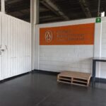 Interior de un almacén con paredes de bloques de cemento. En la pared derecha, se observa un letrero con el logo y el nombre de "Penguin Random House Grupo Editorial" en dos colores. A la izquierda, una puerta cerrada de metal con un candado. En el suelo, hay un palé de madera y una mesa metálica.