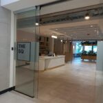 Vista de entrada principal de oficina moderna con puerta corrediza de vidrio. Al fondo se aprecia la recepción y áreas de coworking con plantas decorativas y luz natural.