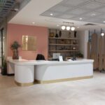 Recepción de oficina elegante con un escritorio curvo de bordes dorados, decoración minimalista y fondo de pared con un letrero. Se observan áreas de trabajo abiertas y salas privadas al fondo.