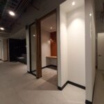 Espacio de trabajo privado o cabina de concentración en un pasillo de oficinas. La cabina tiene una pared de madera, una lámpara colgante y un pequeño escritorio integrado. Al fondo del pasillo se observan sofás de espera.