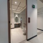 Baño moderno de acceso universal en un edificio corporativo. El baño tiene entrada amplia sin puerta, inodoro a la derecha, lavabo suspendido frente a un espejo ovalado, y paredes cubiertas de azulejos rectangulares.