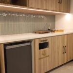 Área de cocina de oficina moderna con muebles de madera clara y un panel de azulejos verticales. Hay un pequeño bote de basura y un gabinete para reciclaje de papel y cartón.