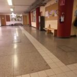 Pasillo amplio dentro de una institución. En el techo se observan varios tubos expuestos. A un lado hay columnas, un tablero de anuncios, un extintor y un banco con una silla. En el fondo se ve una ventanilla de atención y varias puertas. En el piso hay un camino táctil guía para personas con discapacidad visual.