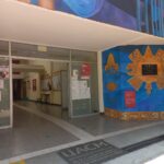 Entrada principal de una universidad. Hay una puerta de vidrio abierta que conduce a un pasillo interior con tableros de anuncios en las paredes. A la derecha, un mural con formas y figuras culturales y una placa. En el suelo, una loseta con el nombre de la universidad y su lema. También hay carteles informativos visibles tanto en la puerta como en las paredes del pasillo.