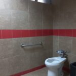 Baño individual con un inodoro sin tapa, un bote de basura a un lado y un tubo metálico horizontal instalado en la pared como barra de apoyo. En la parte superior hay una ventana con rejillas que permite la ventilación e iluminación natural.