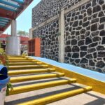 Escaleras que conducen a un edificio con una pared de piedra. Junto a las escaleras, hay una rampa que permite el acceso sin escalones. La fachada del edificio tiene un patrón de piedra en dos tonos. Sobre las escaleras se observan elementos estructurales como pilares y vigas. En primer plano, a la izquierda, hay un bote de basura con la etiqueta "PET".