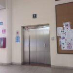 Pasillo de una universidad con paredes lisas y piso de baldosas. Al centro, un elevador con puertas cerradas. Encima del elevador, un letrero indica la salida de emergencia hacia arriba. A la izquierda, hay varios carteles informativos y un extintor montado en la pared. A la derecha, un tablero de corcho con anuncios y carteles, uno de ellos con la palabra "SALUD". En la parte superior derecha, un letrero muestra el texto "Universidad Autónoma" y "Consejo de".