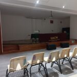 Auditorio vacío con varias filas de sillas orientadas hacia un escenario al frente. Sobre el escenario, una mesa con un mantel que tiene impresas las siglas "UACM" y el texto "Universidad Autónoma de la Ciudad de México". Encima de la mesa hay dos micrófonos en soportes y una computadora portátil. El espacio está iluminado con luces en el techo, y frente al escenario se encuentran altavoces colocados en el suelo.