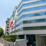 Entrada principal de un edificio con fachada de vidrio, escalinata al frente, barandales metálicos y tres banderas en astas colocadas junto a la banqueta, que también conduce a la misma plaza.