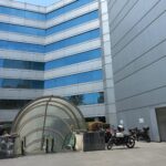 Entrada lateral de un edificio con escaleras eléctricas techadas que conducen al interior de una plaza, con motocicletas estacionadas a un costado.