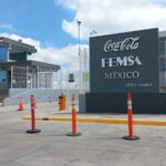 Entrada a una instalación con un letrero que dice "Coca-Cola FEMSA México CEDIS Puebla". Frente a la entrada hay conos de tráfico colocados en el suelo. A la izquierda se encuentra una caseta de seguridad. Al fondo se observa una reja y varios vehículos estacionados.