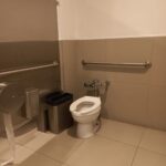 Sanitario individual en un baño público. En el interior hay un escusado, dispensador de papel higiénico y dos botes de basura. En las paredes laterales se encuentran barras de apoyo, el piso y la parte inferior de las paredes están recubiertos con azulejos, mientras que la parte superior de las paredes están lisa.