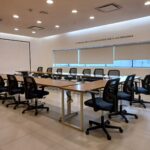 Sala de capacitación con varias sillas dispuestas alrededor de dos mesas alargadas. El espacio se presenta ordenado y limpio, con paredes lisas y piso de loseta. En el techo se observan luminarias y rejillas de ventilación. Uno de los muros cuenta con una ventana grande con persianas, que permite la entrada de luz natural. Sobre esa ventana hay un texto en español que dice: "LA TECNOLOGÍA ES MEJOR CUANDO UNE A LAS PERSONAS".