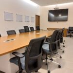 Sala de conferencias con una mesa alargada rodeada por varias sillas de oficina. Hay ocho sillas alrededor de la mesa y dos más colocadas contra la pared del lado derecho. En un extremo de la sala, hay una pantalla plana grande montada en la pared y un proyector suspendido del techo. En la pared izquierda se encuentran seis pizarras montadas. Al fondo se observa una puerta. El piso está recubierto con losetas.