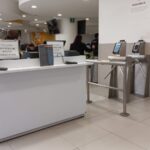 Área de entrada al comedor. Hay un mostrador con dos letreros visibles y un torniquete al lado. Detrás del mostrador, se observan personas sentadas en mesas comiendo. Los letreros sobre el mostrador indican: "LOS POSTRES NO INCLUYEN BEBIDAS NI CAFÉ". En las paredes hay decoraciones con palabras en español como "EXCELENCIA", "ESTILO" y "CALIDEZ".