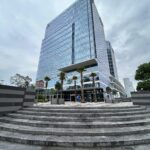 Edificio moderno de varios pisos con fachada de vidrio. Está rodeado por palmeras y otras plantas. En primer plano, hay una escalinata amplia de piedra que conduce a la entrada del edificio. El cielo está cubierto por nubes.