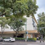 Tienda comercial de dos niveles, con fachada en tonalidades de colores claros. En la planta baja, amplios ventanales muestran productos de alta gama. El entorno está enmarcado por árboles frondosos que rodean la estructura. En una de las esquinas del edificio se ubican un semáforo y señales de tránsito.