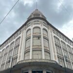 Edificio histórico, destaca en el último nivel una cúpula de azulejos pequeños. En la parte media tres niveles de ventanas arqueadas y detalles arquitectónicos decorados. En la planta baja el acceso principal con el texto "Palacio de Hierro" y ventanas con productos de marca de alta gama.