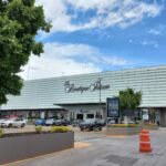 Edificio comercial de diseño moderno, con fachada de paneles de vidrio. En la parte superior se encuentra el nombre "La Boutique Palacio". En la planta baja, el acceso principal está flanqueado por varios automóviles estacionados. El entorno incluye árboles y jardineras que aportan vegetación al espacio.