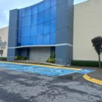 Edificio comercial con fachada de vidrio y el nombre "Outlet Centro" visible en un costado. Al frente hay un área de estacionamiento con espacios designados para personas con discapacidad, señalizados con letreros. La entrada principal cuenta con una cortina metálica cerrada. El entorno incluye árboles pequeños y arbustos.