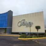 Edificio comercial con diseño moderno. En la parte izquierda, paneles de vidrio cubren varios niveles, mientras que en el lado derecho una pared amplia muestra el letrero "Palacio OUTLET". Al frente hay árboles pequeños, arbustos y un poste de alumbrado. El suelo se ve húmedo, lo que sugiere que ha llovido recientemente.