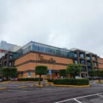 Edificio moderno de varios niveles con fachada que combina paneles lisos y ventanales amplios. En la parte frontal se encuentra el letrero "Palacio de Hierro". Al frente del edificio hay árboles podados, arbustos y un estacionamiento vacío con guarniciones señalizadas. El cielo está cubierto por nubes.