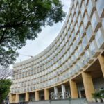Edificio moderno con estructura curva y múltiples ventanas. Al frente se encuentra un letrero con el número "843B" y flechas que indican direcciones hacia "antora" y "VALET". A la izquierda hay árboles, y en el fondo se observa una grúa.