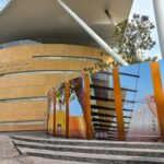 Edificio moderno con fachada curva revestida de piedra. En la parte frontal se encuentra el nombre "antara" en letras estilizadas. Frente al edificio hay paneles verticales con imágenes y textos diversos. Uno muestra a una persona con vestimenta llamativa, otro una estructura arquitectónica, y otro más a una persona saltando. En uno de los paneles se lee el texto "EL PODER DE D." También se observan íconos de redes sociales en la parte inferior de uno de los paneles.