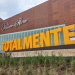 Vista exterior de la tienda departamental El Palacio de Hierro, con un gran letrero amarillo que dice 'TOTALMENTE' al frente y el nombre 'El Palacio de Hierro' en la fachada superior de madera. Se aprecian algunas plantas en primer plano y un edificio alto al fondo.